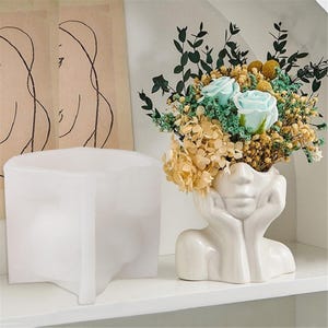 Może przedstawiać: Biały ceramiczny wazon w kształcie twarzy z bukietem kwiatów, w tym niebieskimi różami i żółtymi akcentami. Obok wazonu znajduje się biała silikonowa forma twarzy. Wazon ma dłonie podtrzymujące twarz.