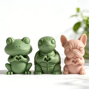 Op de afbeelding: Drie decoratieve beeldjes: een groene kikker, een groene schildpad en een roze Franse bulldog. Elke figuur houdt zijn handen in een hartvorm. De kikker en de schildpad hebben een matte afwerking, terwijl de bulldog een gladde textuur heeft.