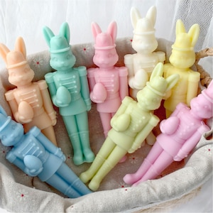 Puede incluir: Una colección de figuras de conejitos en colores pastel en una cesta tejida. Cada conejito viste un uniforme de estilo militar y sostiene un huevo. Los colores incluyen rosa, azul, amarillo, verde y melocotón. Las figuras son probablemente artículos decorativos.