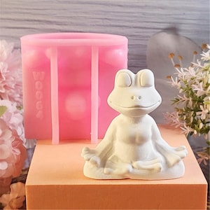 May include: A white frog figurine in a meditative pose, with arms resting on its knees. A pink silicone mold with the word "Booca" is behind the frog. The frog is on a peach-colored surface.