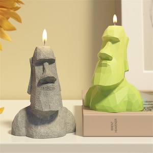 Puede incluir: Dos velas encendidas con forma de estatuas de la Isla de Pascua. Una es gris y texturizada, la otra es verde lima y geométrica. Las velas están sobre una superficie blanca, con un libro visible en el fondo.