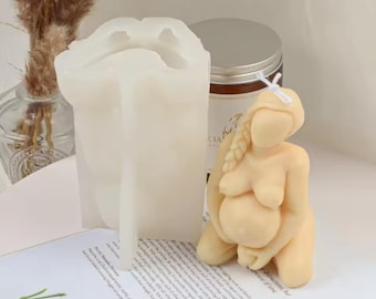 May include: A light beige candle in the shape of a pregnant person kneeling, with a long braid. Beside it is a white silicone mold in the shape of a pregnant person. A jar with a silver lid is in the background.