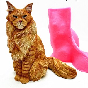 Può includere: Una statuetta di gatto arancione e marrone dettagliata con una lunga coda e pelliccia soffice. Il gatto è seduto in posizione eretta. Uno stampo rosa brillante è accanto al gatto.