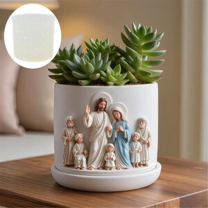 May include: White ceramic planter with a relief of religious figures, including Jesus, Mary, and children. The planter holds green succulents and sits on a matching saucer. A small, decorative, white, cylindrical object is in the upper left corner.