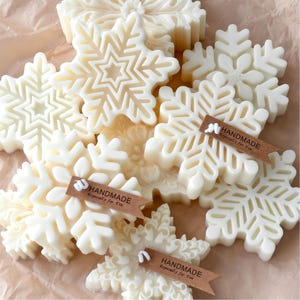 May include: A selection of white snowflake-shaped soaps. Each soap features detailed patterns, mimicking real snowflakes. Some soaps have small brown tags that read "HANDMADE Especially for You". The soaps are arranged on a light brown surface.