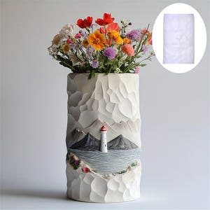 Può includere: Un vaso di ceramica bianca con un design scolpito e strutturato con una scena di faro. Il vaso contiene una vivace composizione di fiori colorati. Il faro ha una parte superiore rossa e il vaso è alto circa 30 cm.