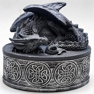 May include: A detailed, dark gray dragon figurine atop a round base. The dragon has textured scales, wings, and a detailed head. The base features a Celtic knot design around its perimeter, creating a fantasy-themed decorative piece.