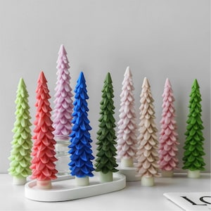 Puede incluir: Una colección de velas decorativas en forma de árbol de Navidad, coloridas. Las velas son de varios tonos de verde, rojo, rosa, azul y blanco. Están dispuestas sobre una bandeja blanca y un soporte de vidrio, creando una exhibición festiva.