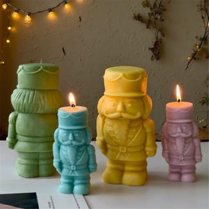 Puede incluir: Cuatro velas encendidas con forma de cascanueces en verde, azul, amarillo y rosa. Las velas tienen uniformes y sombreros detallados. Están dispuestas en fila sobre un fondo neutro, con luces.