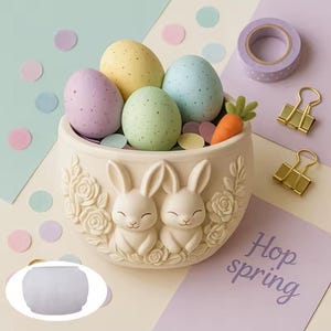 May include: A cream-colored ceramic Easter egg holder with two bunny figures and floral relief. The holder contains speckled pastel eggs, a felt carrot, and confetti. A roll of purple polka dot tape and gold binder clips are also present.