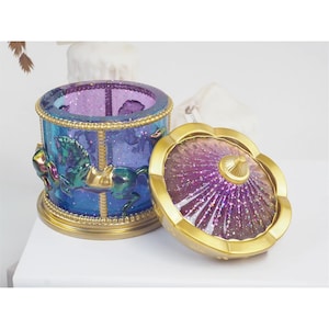 May include: A carousel-themed trinket box with a purple and blue glass lid and base. The lid features a gold rim and a small gold dome in the center. The base features a gold rim and a scene of horses on a carousel.