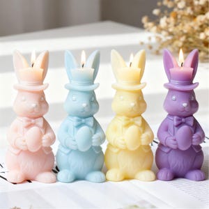May include: Four pastel-coloured Easter bunny candles: pink, blue, yellow, and purple. Each bunny wears a top hat and holds an egg. The candles have lit wicks.