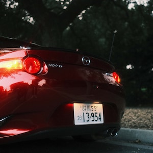 Może przedstawiać: Widok z tyłu czerwonego samochodu sportowego Mazda MX-5 z podświetlonymi tylnymi światłami. Widoczna japońska tablica rejestracyjna. Samoch&oacute;d zaparkowany na drodze, w tle ciemne drzewo.
