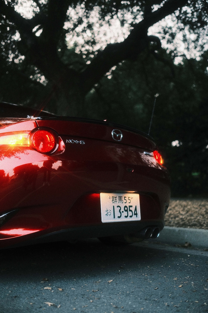 Może przedstawiać: Widok z tyłu czerwonego samochodu sportowego Mazda MX-5 z podświetlonymi tylnymi światłami. Widoczna japońska tablica rejestracyjna. Samoch&oacute;d zaparkowany na drodze, w tle ciemne drzewo.