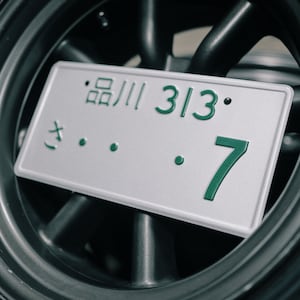 Może przedstawiać: Zbliżenie japońskiej tablicy rejestracyjnej z zielonym tekstem "品川 313 7" na srebrnym tle. Tablica umieszczona na tle ciemnoszarego koła.