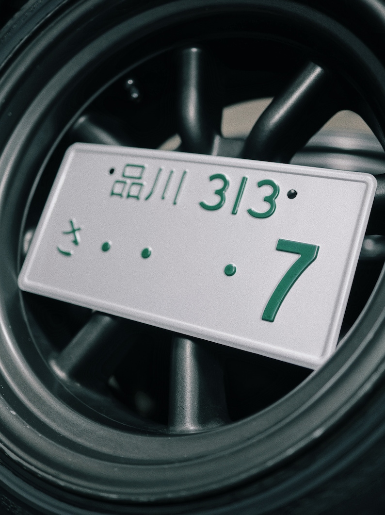 Może przedstawiać: Zbliżenie japońskiej tablicy rejestracyjnej z zielonym tekstem "品川 313 7" na srebrnym tle. Tablica umieszczona na tle ciemnoszarego koła.