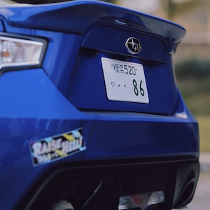 Może przedstawiać: Widok z tyłu niebieskiego samochodu sportowego Subaru. Samoch&oacute;d ma spojler, tablicę rejestracyjną z japońskimi znakami i numerem 86 oraz naklejkę z napisem "RAISE HELL". Widoczna jest rura wydechowa.