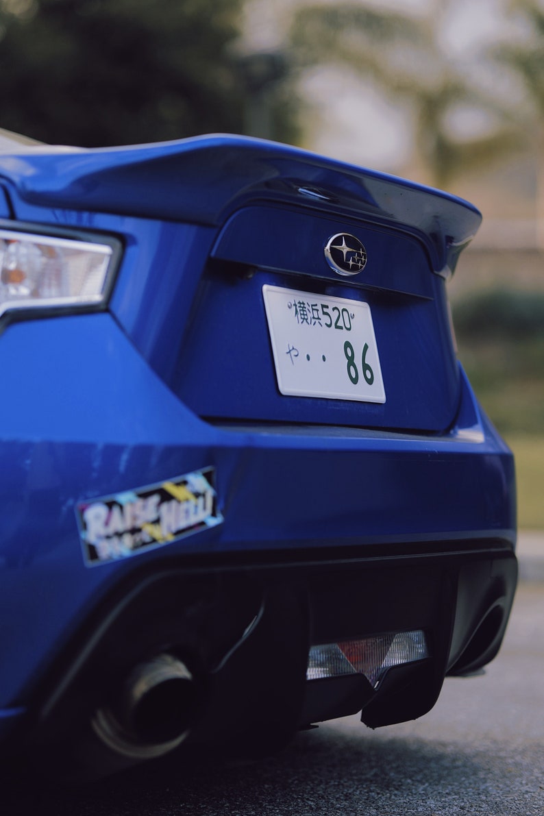 Może przedstawiać: Widok z tyłu niebieskiego samochodu sportowego Subaru. Samoch&oacute;d ma spojler, tablicę rejestracyjną z japońskimi znakami i numerem 86 oraz naklejkę z napisem "RAISE HELL". Widoczna jest rura wydechowa.