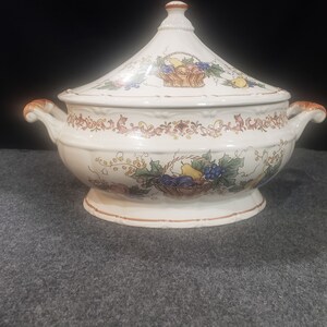 May include: A white ceramic serving dish with a lid. The dish is decorated with a floral pattern and a basket of fruit. The lid has a knob in the shape of a fruit.