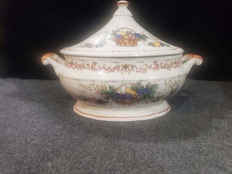 May include: A white ceramic serving dish with a lid. The dish is decorated with a floral pattern and a basket of fruit. The lid has a knob in the shape of a fruit.