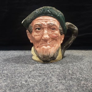 Puede incluir: Una taza de cerámica con forma de cabeza de hombre. La taza está pintada para parecer un hombre mayor con un sombrero verde, barba marrón y una sonrisa traviesa.