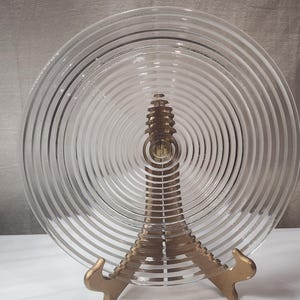 Puede incluir: Plato de vidrio transparente con crestas circulares concéntricas, exhibido en un soporte de metal dorado. El diseño del plato crea un efecto de espiral. El soporte tiene una forma decorativa y estilizada.