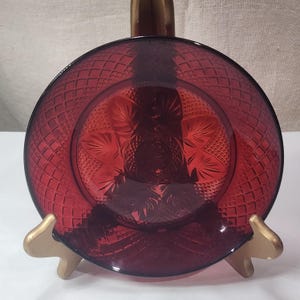 Op de afbeelding: Een robijnrode glazen schotel met een ruitpatroon langs de rand en een bloemmotief in het midden. De schotel wordt gepresenteerd op een goudkleurige standaard, die de ingewikkelde details en vintage charme benadrukt. Diameter ongeveer 20 cm.