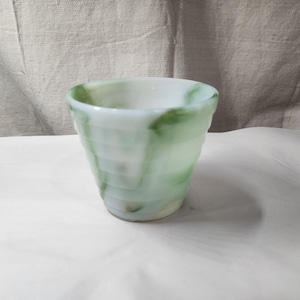 Op de afbeelding: Een kleine, ronde, geribbelde beker van wit melkglas met wervelende groene patronen over de gehele buitenkant. De beker heeft een brede, open bovenkant en een taps toelopende voet, met een doorschijnend, gemarmerd uiterlijk met horizontale ribbels.