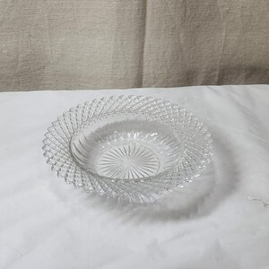 May include: Clear glass bowl with a decorative, diamond-patterned rim. The bowl has a circular shape with a textured interior and a wide, scalloped edge. The bowl is sitting on a white surface.