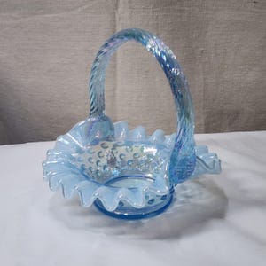 Op de afbeelding: Een lichtblauwe glazen mand met een gekrulde rand en een gedraaide handgreep. De mand is versierd met kleine witte stippen.