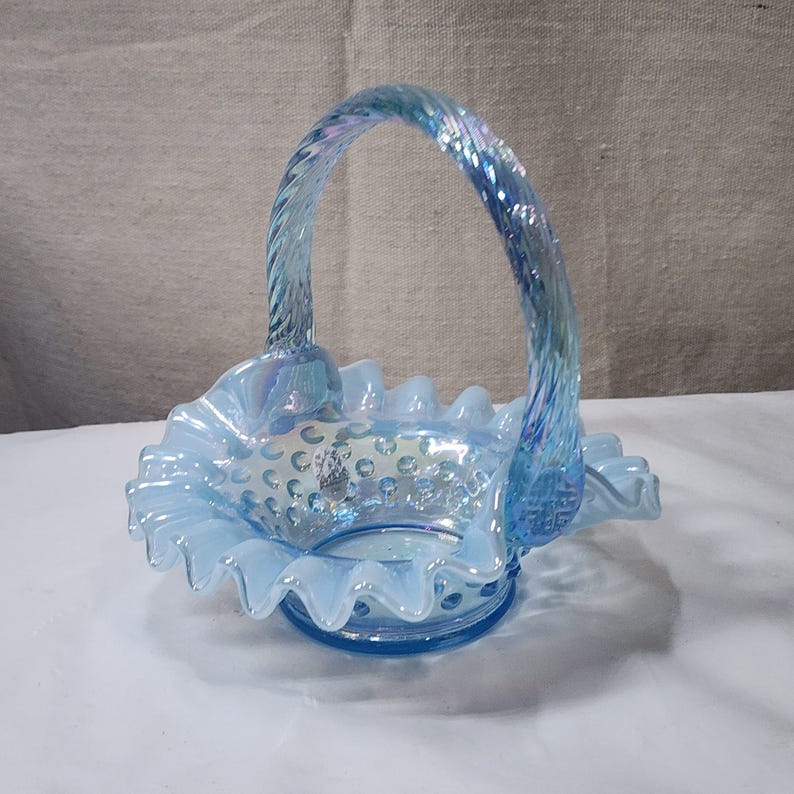 Op de afbeelding: Een lichtblauwe glazen mand met een gekrulde rand en een gedraaide handgreep. De mand is versierd met kleine witte stippen.