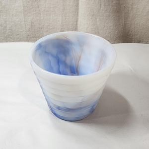 Op de afbeelding: Een kom van wit melkglas met een wervelend blauw gemarmerd patroon over het hele lichaam. De buitenkant van de kom heeft een reeks horizontale ribbels of ringen die rond de omtrek lopen, waardoor een getextureerd oppervlak ontstaat.