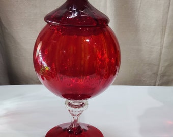 May include: A red glass candy jar with a round body and a domed lid. The jar sits on a clear glass pedestal with a red base.