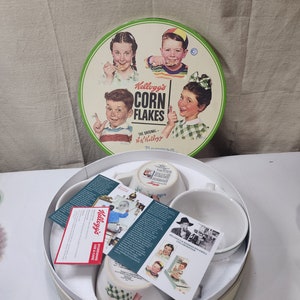 Op de afbeelding: Een witte doos met een groen deksel met daarin vier witte keramische kommen met een Kellogg's Corn Flakes-thema. De kommen bevatten illustraties van kinderen die van de ontbijtgranen genieten. Het deksel van de doos heeft een vintage Kellogg's Corn Flakes-reclame met vier kinderen.