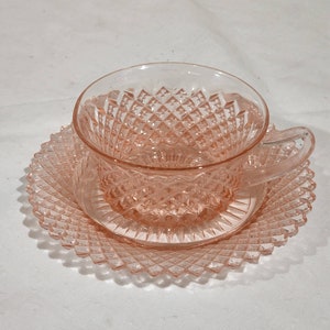 Può includere: Un set da tè in vetro rosa con un motivo a diamante. La tazza ha un manico e il piattino è rotondo.