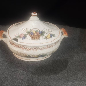 May include: A white ceramic serving dish with a lid, featuring a floral design with fruit and a basket of fruit. The dish has a brown rim and two brown handles.