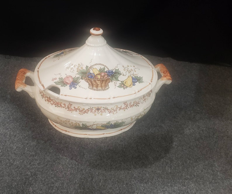 May include: A white ceramic serving dish with a lid, featuring a floral design with fruit and a basket of fruit. The dish has a brown rim and two brown handles.