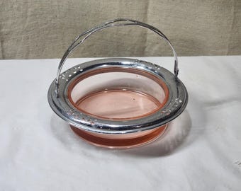 Pode incluir: Uma cesta de vidro rosa vintage com aro e alça de metal prateado. A borda apresenta um design floral decorativo. A cesta é provavelmente usada para servir ou exibir.