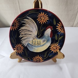 May include: A blue ceramic plate featuring a hand-painted design of a rooster with a red comb and white feathers. The rooster is surrounded by sunflowers with yellow petals and brown centres. The plate is on a gold stand.