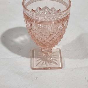 May include: A pink glass goblet with a square base and a diamond pattern. The goblet is made of clear glass with a pink tint.