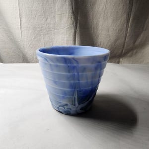 Op de afbeelding: Een taps toelopende, cilindrische plantenbak met een geribbelde buitenkant. De pot is gemaakt van gemarmerd glas met een mix van lichtblauwe, donkerblauwe en witte wervelende patronen over het gehele oppervlak.