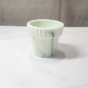 Op de afbeelding: Een kleine, wit en groen gemarmerde glazen plantenbak. De plantenbak heeft een geribbelde bovenkant en een cilindrische vorm. De binnenkant is lichtgroen van kleur. De plantenbak staat op een wit oppervlak.
