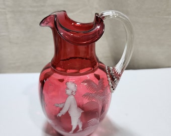 Puede incluir: Una pequeña jarra de vidrio roja con un asa de vidrio transparente. La jarra tiene un diseño grabado de una persona y una palmera.