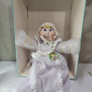 May include: A Cabbage Patch Kid doll wearing a white dress with lace trim and a yellow bonnet. The doll has blonde hair, blue eyes, and a pink bow on its chest. The doll is sitting in a white box with a blue and white striped lining.