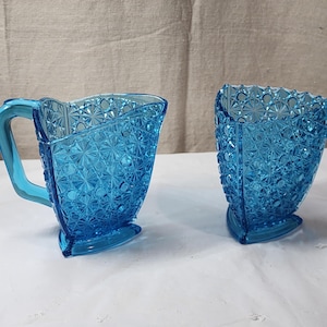 Könnte beinhalten: Zwei Teile Vintage-Geschirr aus blauem Glas. Eines ist ein kleiner Krug mit Henkel, das andere ein passender Becher. Beide weisen ein Diamanten- und Sternmuster auf, ideal zum Servieren von Getränken.