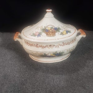 May include: A white ceramic serving dish with a lid. The lid has a floral design with a basket of fruit. The dish has a brown rim and two handles.