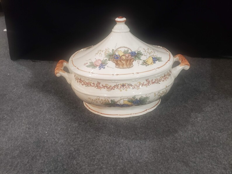 May include: A white ceramic serving dish with a lid. The lid has a floral design with a basket of fruit. The dish has a brown rim and two handles.