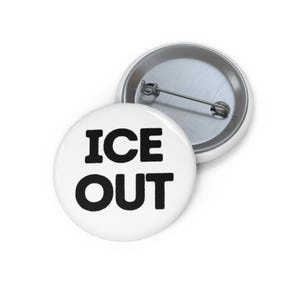 Può includere: Due distintivi rotondi bianchi con le parole "ICE OUT" in grassetto nero. Un distintivo è a fuoco, mentre l'altro è leggermente sfocato, mostrando la spilla sul retro. I distintivi sono su una superficie bianca.