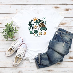 May include: White t-shirt with a circular graphic of a hiker, mountains, tent, and other camping elements. The shirt is paired with blue jeans and white canvas sneakers.