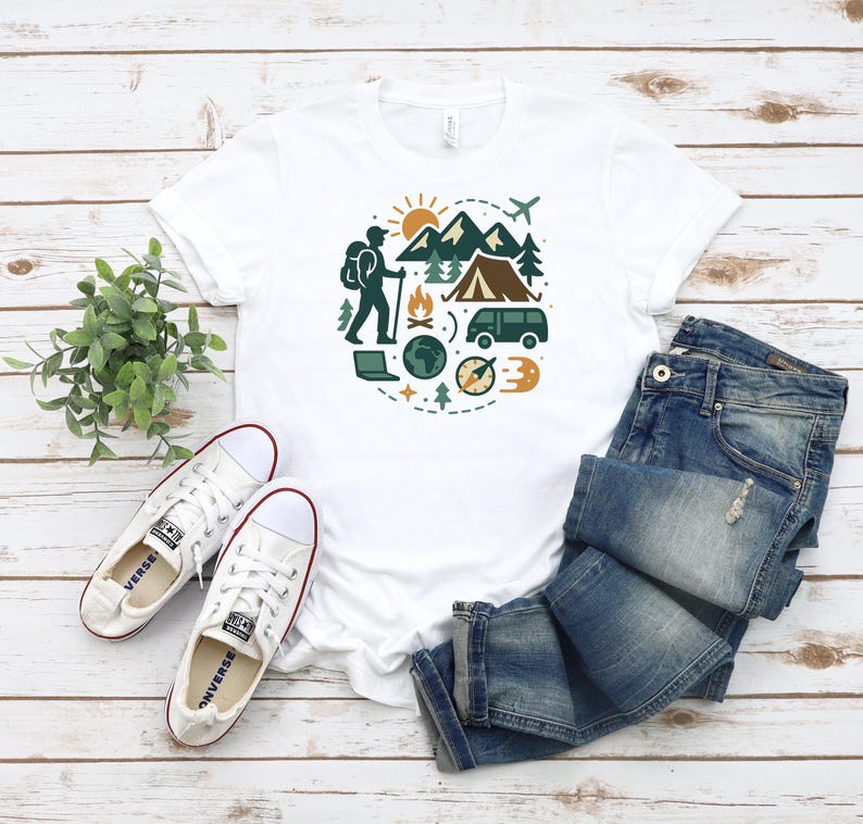 May include: White t-shirt with a circular graphic of a hiker, mountains, tent, and other camping elements. The shirt is paired with blue jeans and white canvas sneakers.